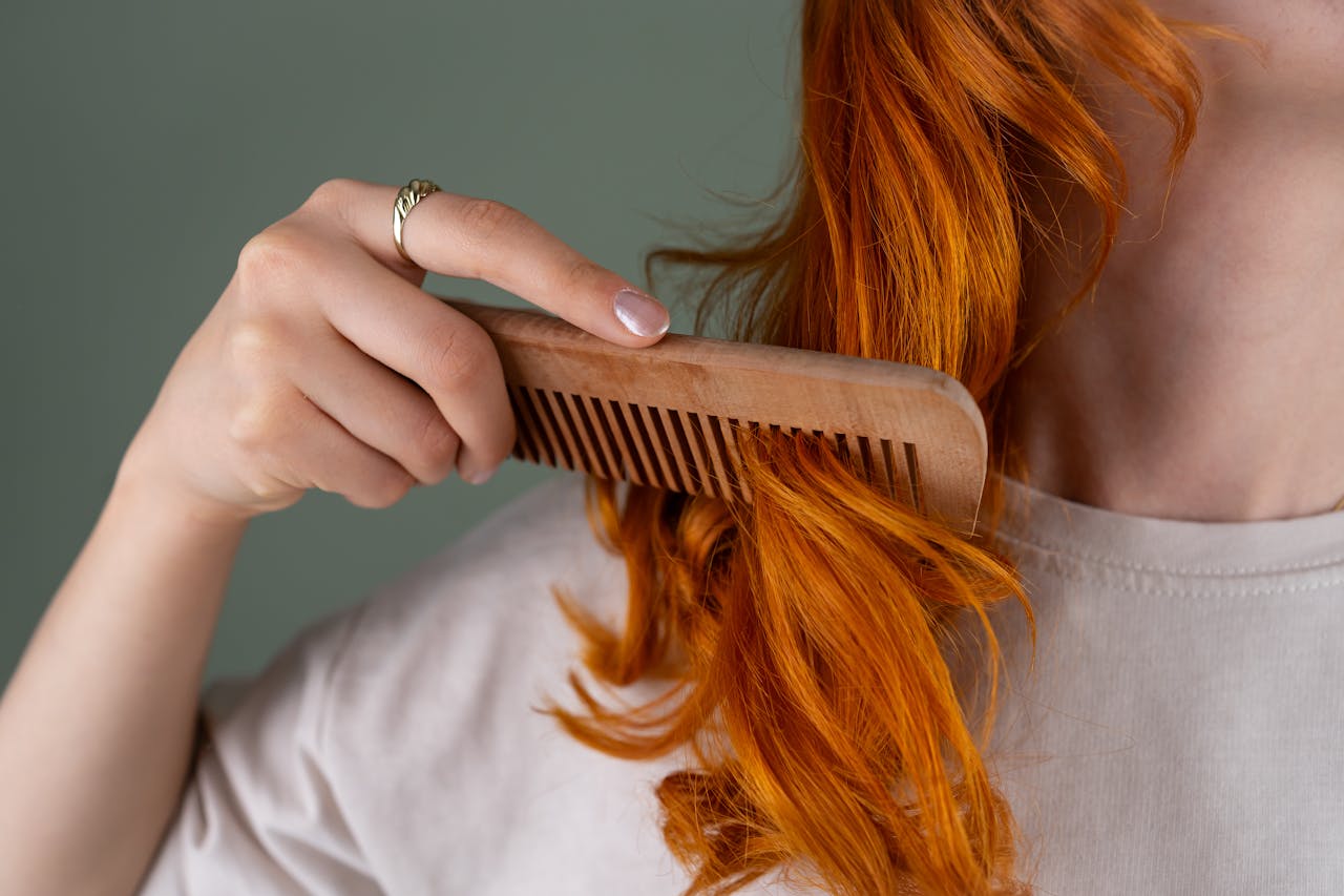 hero-img-01 Close-up of a woman combing her red curly hair using a wooden comb against a green background.